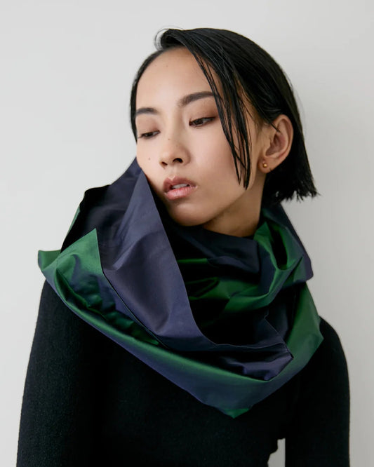 A model with short black hair is wearing a dark navy and forest green satin-like scarf wrapped around her neck over a black long-sleeve top, set against a plain light gray background.