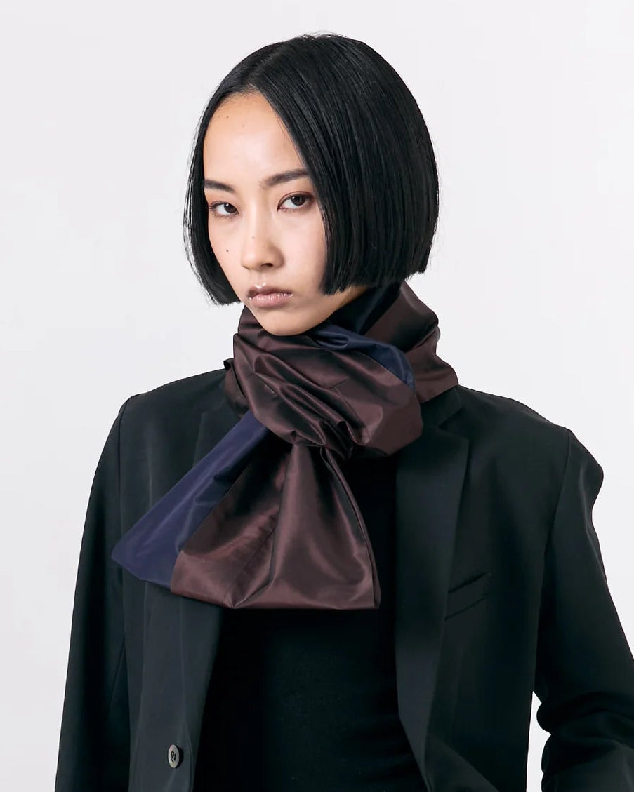 A model wearing a black blazer, black trousers, and a dark brown satin bow tie or scarf, photographed against a plain white studio background.